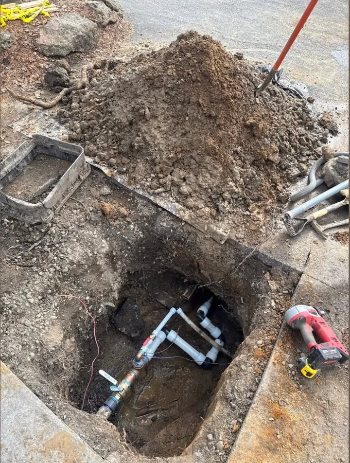 Excavation beside a sidewalk exposing underground plumbing pipes and a brass valve for repair